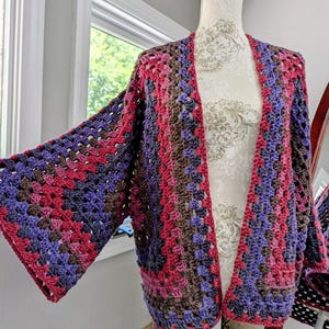 May include: A handmade, open-front crochet cardigan in a granny square pattern. The color palette includes shades of pink, purple, and brown. The cardigan is displayed on a mannequin, showcasing its loose fit and wide sleeves. This garment is a cozy and stylish addition to any wardrobe.