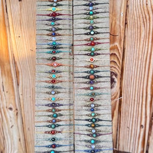 Op de afbeelding: Een display van kleurrijke armbandjes van touw met verschillende edelsteen kralen. De armbandjes zijn gerangschikt in rijen op een beige achtergrond.