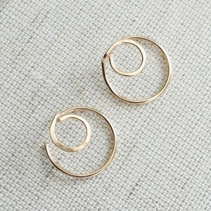 May include: Two gold-colored spiral earrings. Each earring features a larger outer circle with a smaller inner circle, creating a layered, open design. The earrings are displayed on a textured, light-colored surface.