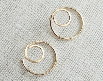 May include: Two gold-colored spiral earrings. Each earring features a larger outer circle with a smaller inner circle, creating a layered, open design. The earrings are displayed on a textured, light-colored surface.