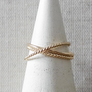 May include: Two gold-colored rings with a crisscross design are displayed on a white, cone-shaped ring holder. The rings have a twisted rope-like texture. The ring holder is made of a textured, matte material.