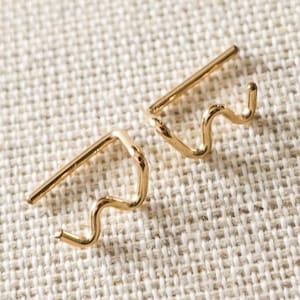 May include: A pair of gold-colored earrings. Each earring features a straight bar with a wavy design extending from it. The earrings are displayed on a textured, off-white surface with a blurred orange background.