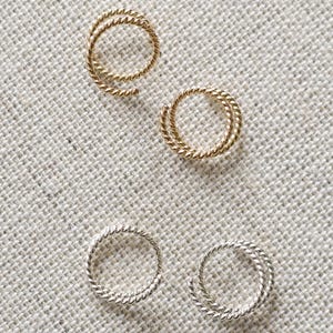 May include: Close-up of three pairs of earrings on a textured, off-white surface. Two pairs are circular, one in gold and two in silver. The third pair is a spiral design in gold. The background is a mix of off-white and tan.