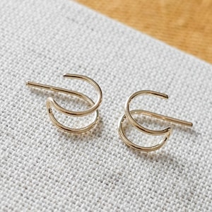 May include: A pair of gold-colored hoop earrings. Each earring features a double-hoop design with a post for insertion. The earrings are displayed on a textured, off-white surface with a yellow and tan background.
