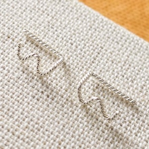 May include: A pair of silver-toned ear climbers. Each earring features a wavy design connected to a straight bar. The earrings are displayed on a textured, off-white surface against a tan background.