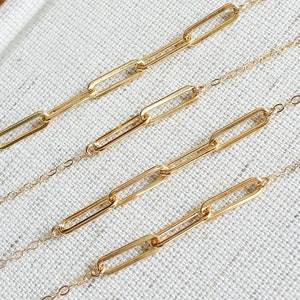 Puede incluir: Cuatro delicados collares de cadena dorada con un diseño de eslabones de clip. Los collares se exhiben sobre una superficie texturizada de color blanquecino. Las cadenas son finas y los eslabones son alargados.