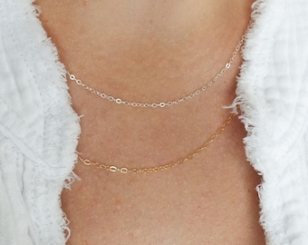 May include: A close-up of a person wearing two delicate necklaces, one silver and one gold, layered on top of each other.