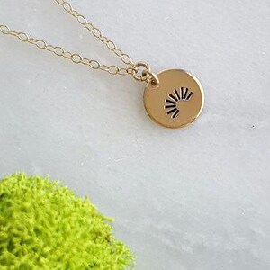 May include: A gold necklace with a small, round pendant featuring a sunburst design. The necklace is on a white surface with green moss.