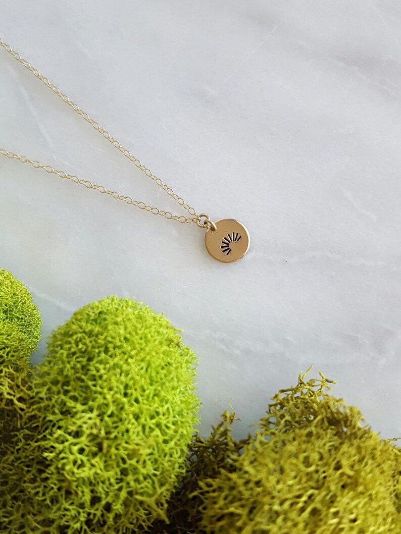 May include: A gold necklace with a small, round pendant featuring a sunburst design. The necklace is on a white surface with green moss.