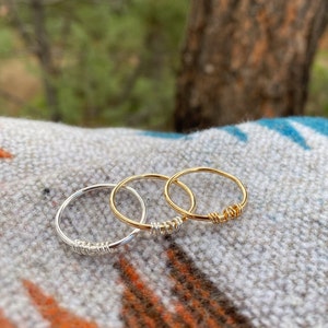 May include: Three simple wire rings, one silver and two gold, stacked on a patterned fabric. The rings are made of thin wire and have a simple, minimalist design.