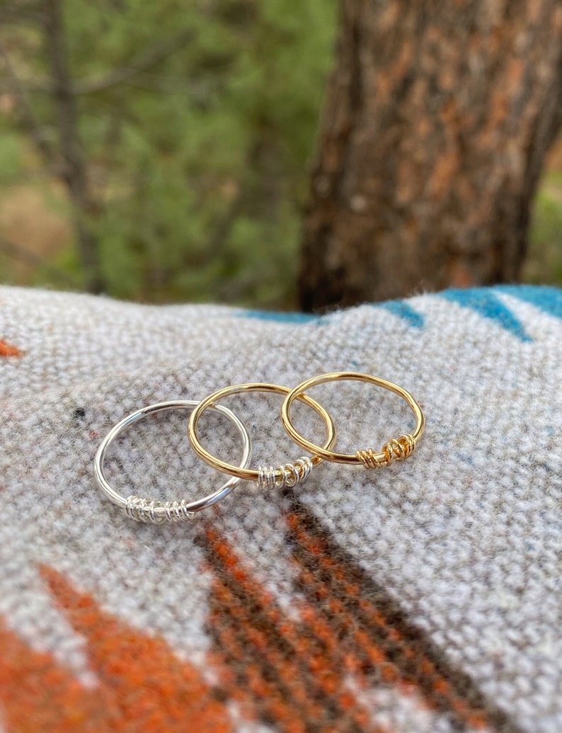 May include: Three simple wire rings, one silver and two gold, stacked on a patterned fabric. The rings are made of thin wire and have a simple, minimalist design.