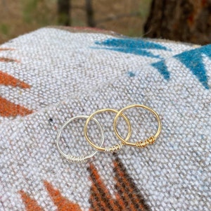 May include: Three simple wire rings, one silver and two gold, are stacked on a patterned fabric surface.