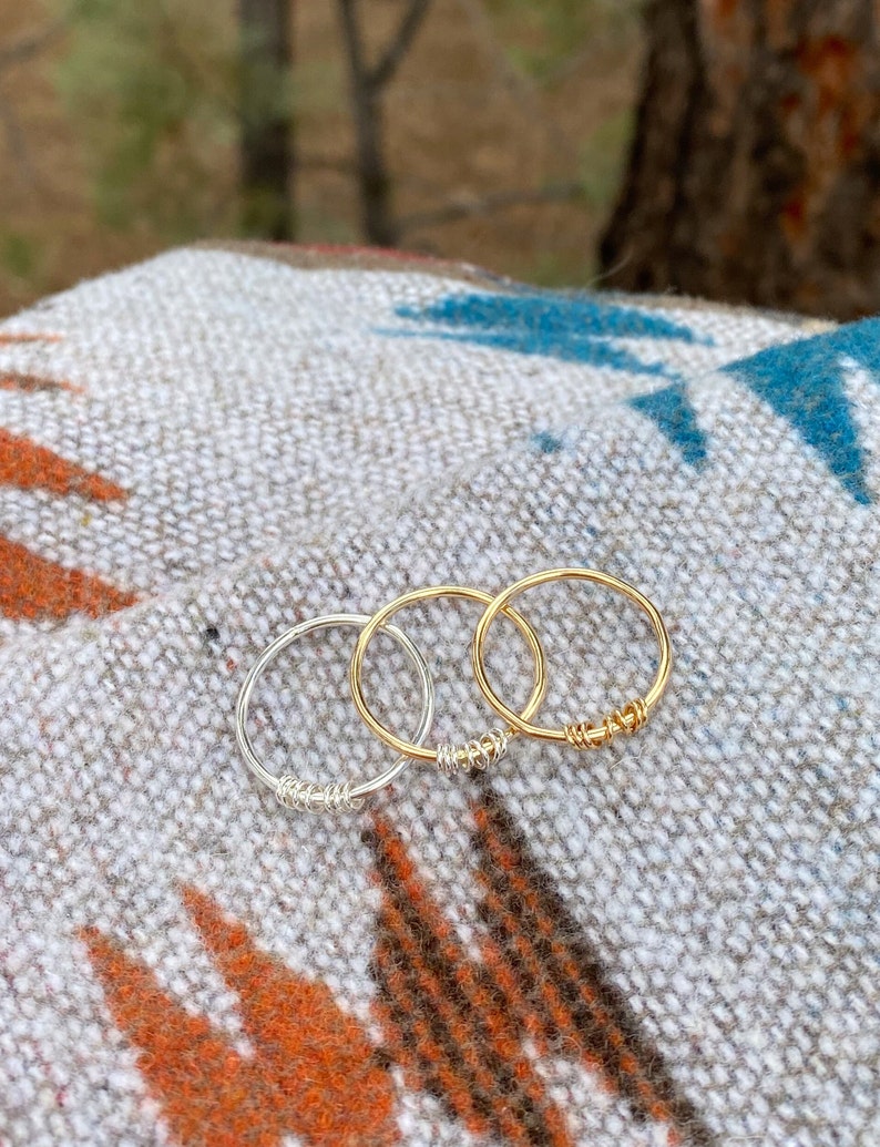 May include: Three simple wire rings, one silver and two gold, are stacked on a patterned fabric surface.