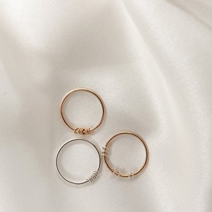 May include: Three simple rings, two gold and one silver, with twisted wire details. The rings are arranged on a white fabric background.