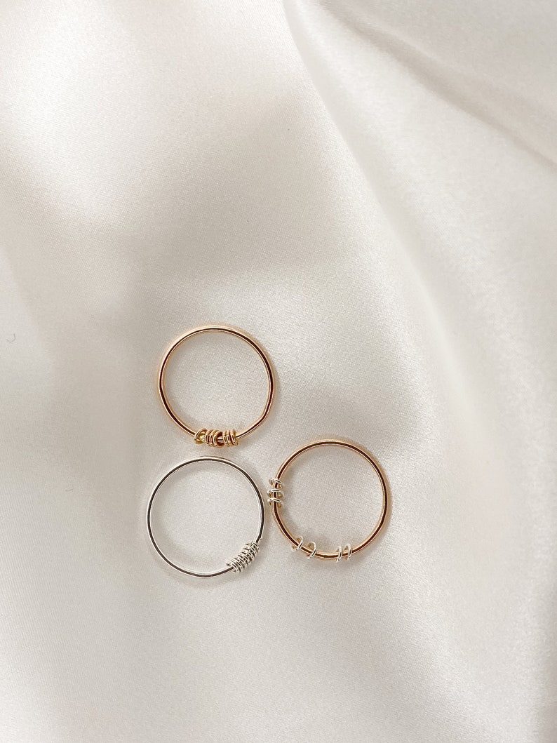 May include: Three simple rings, two gold and one silver, with twisted wire details. The rings are arranged on a white fabric background.