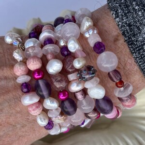 Könnte beinhalten: Eine Sammlung von Armbändern mit Perlen in Rosa-, Lila- und Weißtönen. Die Armbänder bestehen aus Perlen, Rosenquarz und Amethyst. Die Armbänder sind gestapelt und erzeugen einen mehrschichtigen Look. Die Perlen werden in Zentimetern gemessen.