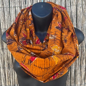 Può includere: Una sciarpa infinita marrone con un motivo batik in tonalità arancio, rosso e giallo. La sciarpa è realizzata in un tessuto morbido e confortevole ed è perfetta per aggiungere un tocco di stile a qualsiasi outfit.