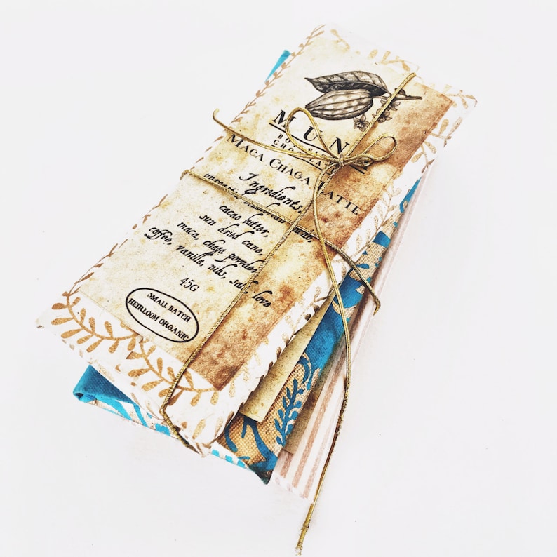 Puede incluir: Una barra de chocolate org&aacute;nico envuelta en papel marr&oacute;n con una cinta dorada. La etiqueta dice "Maca Chaga Latte" y enumera los ingredientes: manteca de cacao, ca&ntilde;a de az&uacute;car seca, polvo de maca chaga, caf&eacute;, vainilla, niks, sal, amor. La barra pesa 45 g y es un producto org&aacute;nico artesanal de peque&ntilde;o lote.