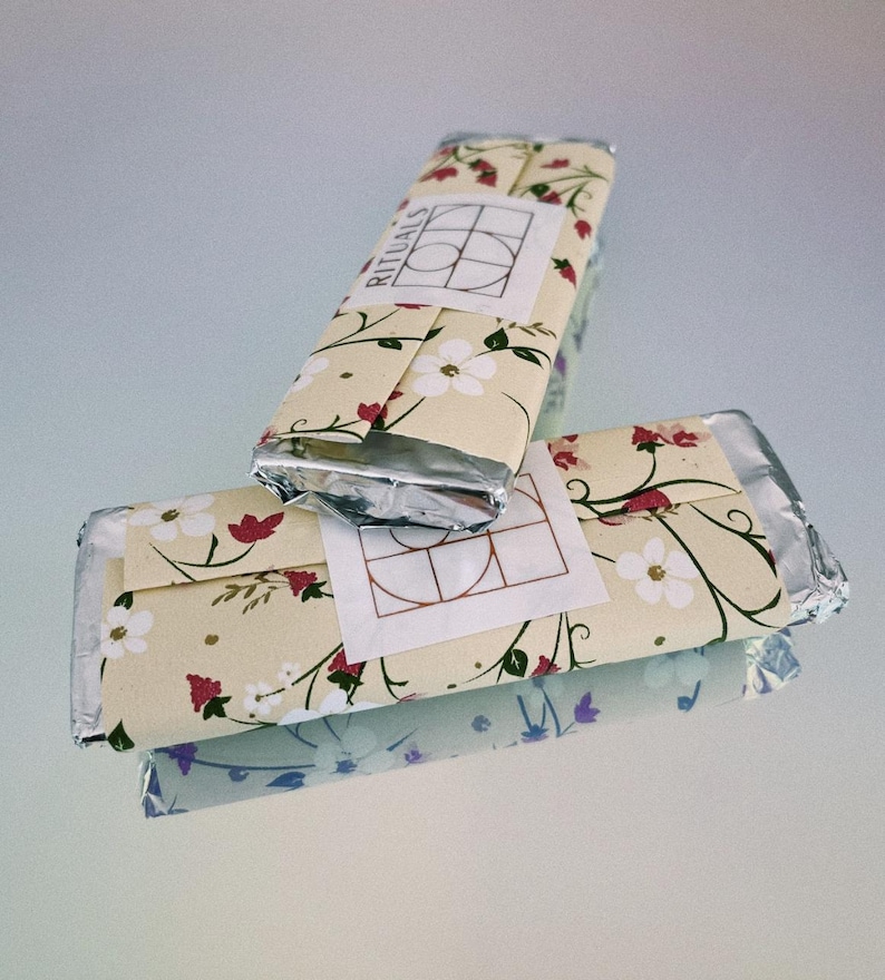 Puede incluir: Dos barras de chocolate envueltas en papel de aluminio plateado con un patr&oacute;n floral y la palabra "RITUALS" impresa en la barra superior.