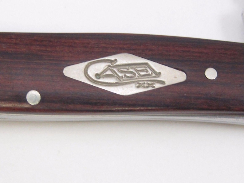 Puede incluir: Primer plano del mango de una navaja de bolsillo con un acabado de veta de madera oscura. Un emblema plateado con la palabra "CASE" est&aacute; incrustado en el mango. Se ven dos remaches plateados.