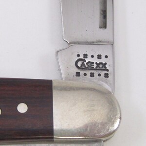 Puede incluir: Primer plano de una navaja de bolsillo parcialmente abierta. El cuchillo tiene un mango de madera marr&oacute;n oscuro con una virola plateada y una hoja de acero inoxidable. La hoja est&aacute; marcada con el texto "CASE XX".