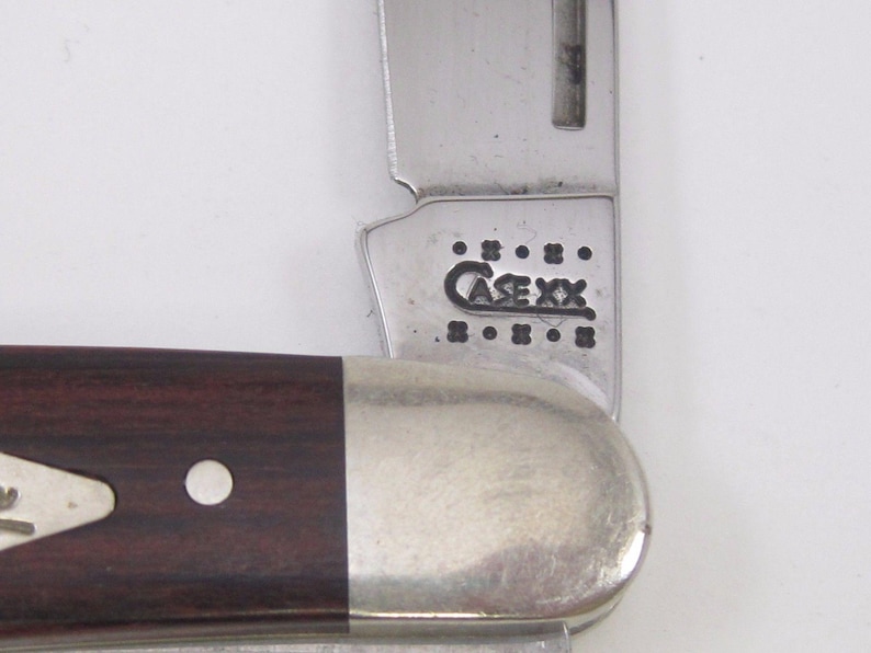 Puede incluir: Primer plano de una navaja de bolsillo parcialmente abierta. El cuchillo tiene un mango de madera marr&oacute;n oscuro con una virola plateada y una hoja de acero inoxidable. La hoja est&aacute; marcada con el texto "CASE XX".