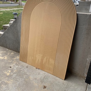May include: A large, unfinished, light brown wood archway with a rounded top and multiple grooves. The archway is leaning against a concrete wall.