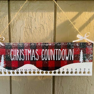 May include: A red and black buffalo plaid Christmas countdown board with white snowflakes and trees. The board has 24 holes for hanging ornaments or other decorations. The text "CHRISTMAS COUNTDOWN" is printed on the board.