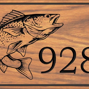 Op de afbeelding: Houten plaquette met een gedetailleerde gravure van een zwarte vis die omhoog springt, tegen een lichte houtnerf achtergrond. Het nummer "928" staat eronder in een vetgedrukte, zwarte letter. De plaquette heeft een zwarte rand.