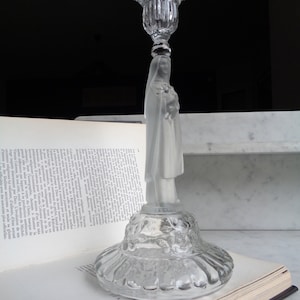 May include: A clear glass candle holder featuring a frosted glass figurine of a woman. The base of the candle holder has a decorative floral design.