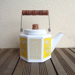 Może przedstawiać: Biały emaliowany czajnik z żółtym i białym wzorem kwiatowym i drewnianym uchwytem. Czajnik ma geometryczny wzór po bokach.