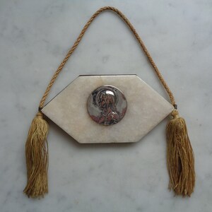 May include: A white marble plaque with a silver-toned image of a woman's face in a circle. The plaque is attached to a beige cord with tassels on each end.
