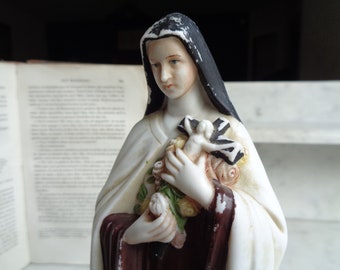 Puede incluir: Figurilla de porcelana blanca de una mujer con una túnica blanca y negra que sostiene un ramo de rosas blancas y una pequeña figura de una persona en una cruz.