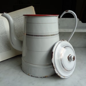 Może przedstawiać: Biały emaliowany dzbanek do kawy z czerwonym rantem i złotymi paskami. Dzbanek ma pokrywkę z gałką na górze. Dzbanek stoi na białym tle.