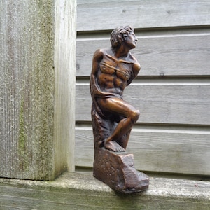 Könnte beinhalten: Eine Bronzeskulptur eines nackten Mannes, der auf einem Felsen steht. Die Figur ist mit einem erhobenen Arm und dem anderen Arm, der am Ellbogen gebeugt ist, dargestellt. Die Skulptur ist detailliert und realistisch.