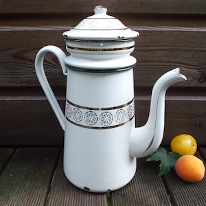 May include: White enamel coffee pot with gold trim and a decorative band of wheel-like designs. The pot has a long spout and a handle. Two apricots and a lemon are next to the pot.