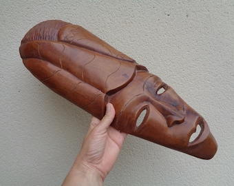 Pode incluir: Uma máscara de madeira com um rosto esculpido e uma forma longa e estreita. A máscara é castanha e tem uma superfície texturizada.