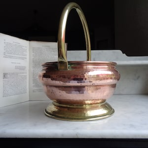 Puede incluir: Un cubo de hielo de cobre martillado y latón con un asa de latón. El cubo tiene una forma redondeada y una base ancha.