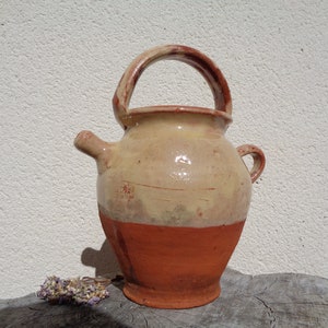 Puede incluir: Jarra de cerámica rústica hecha a mano, con asa y pico vertedor. La jarra presenta un diseño bicolor, con una mitad superior beige claro y una mitad inferior marrón rojizo. Un pequeño arreglo floral seco está delante.