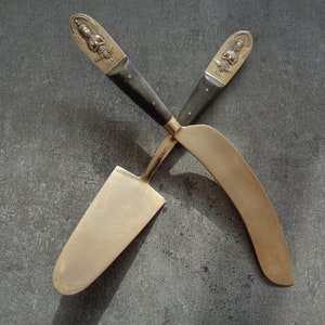 May include: Two ornate brass serving utensils with black wooden handles. The utensils are crossed and feature a decorative design of a seated figure on the handle.