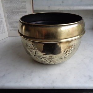 May include: A brass planter featuring a decorative grapevine design. The planter has a dark brown interior and a slightly rounded shape.