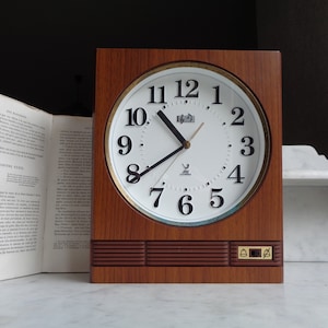 May include: A vintage wooden clock with a white face and black numbers. The clock has a gold trim around the face and a small bell on the side. The clock is set to 10:10.