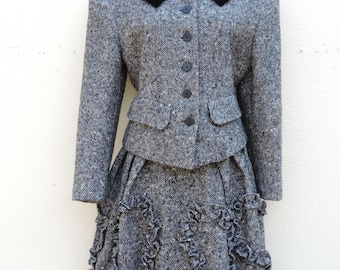 May include: A black and white tweed skirt suit with a black velvet collar. The skirt has a ruffled hem.