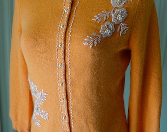 Puede incluir: Un cárdigan de punto naranja con cuello redondo y mangas tres cuartos. La tapeta frontal está adornada con botones blancos tipo perla y abalorios blancos detallados. Adornos florales de cuentas decoran la zona del pecho.