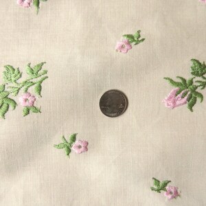 May include: White fabric with a delicate floral pattern embroidered in pink and green. The fabric has a subtle texture and a soft, light feel.