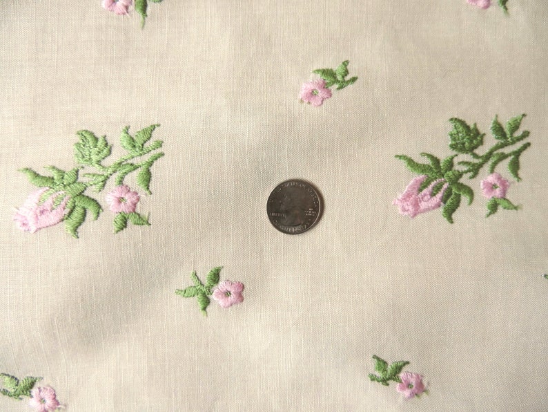 May include: White fabric with a delicate floral pattern embroidered in pink and green. The fabric has a subtle texture and a soft, light feel.