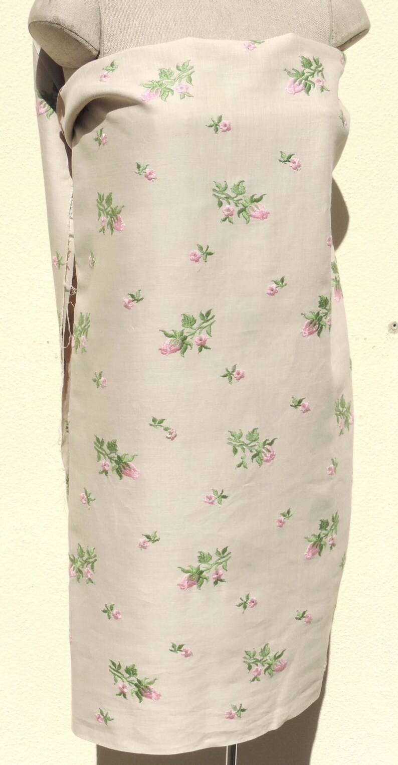 May include: A beige fabric with a delicate floral pattern. The fabric is embroidered with small pink roses and green leaves.