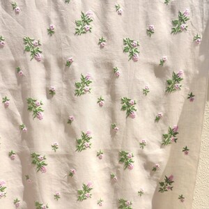 May include: White fabric with a delicate floral pattern embroidered in pink and green. The fabric has a subtle texture and a soft, light feel.