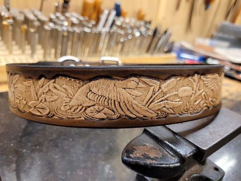 Pode incluir: Um cinto de couro castanho com um design em relevo de um p&aacute;ssaro num p&acirc;ntano. O design &eacute; de cor castanho claro e apresenta juncos e l&iacute;rios-de-&aacute;gua.