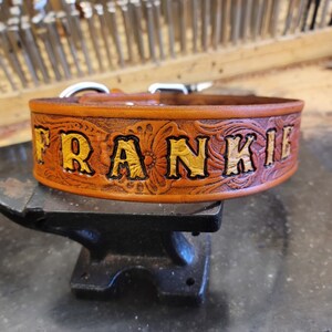 May include: A brown leather dog collar with a floral design and the name "FRANKIE" embossed in gold lettering.